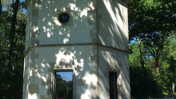 Une petite construction carrée en pierre, dotée d’une fenêtre circulaire et d’une porte, et entourée d’arbres.