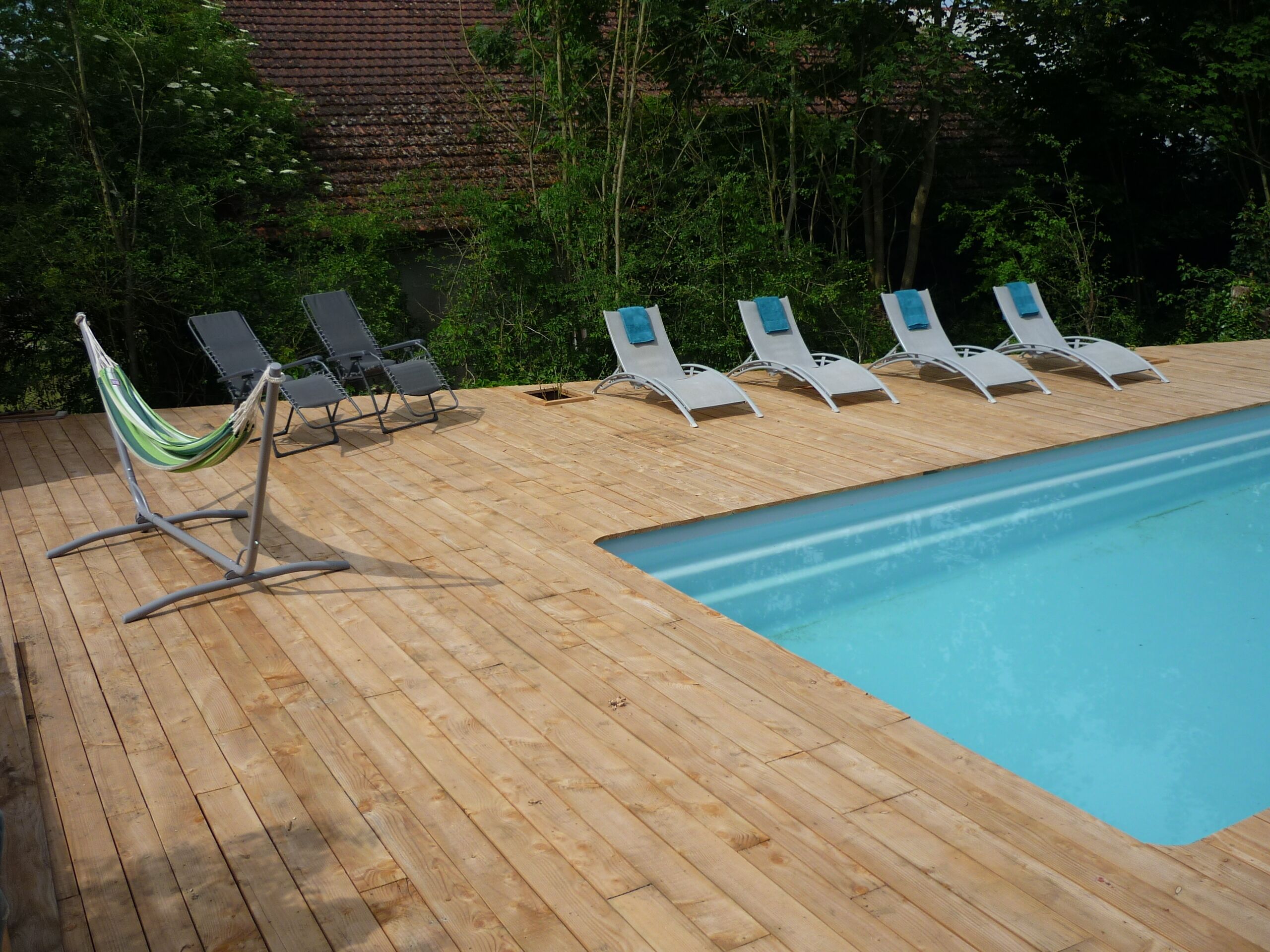 Un terrazzo in legno con piscina, circondato da alberi e un edificio in mattoni sullo sfondo.