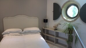 A bedroom with a bed, a nightstand, a lamp, a shelf with decorative items, and a potted plant.
