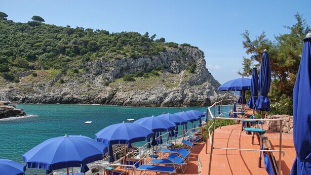 A coastal terrace with blue umbrellas and sun loungers overlooking turquoise waters and a rocky cliff.