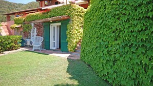 A well-maintained garden with a neatly trimmed hedge, a patio area with a white chair, and a building with green shutters and a large umbrella.