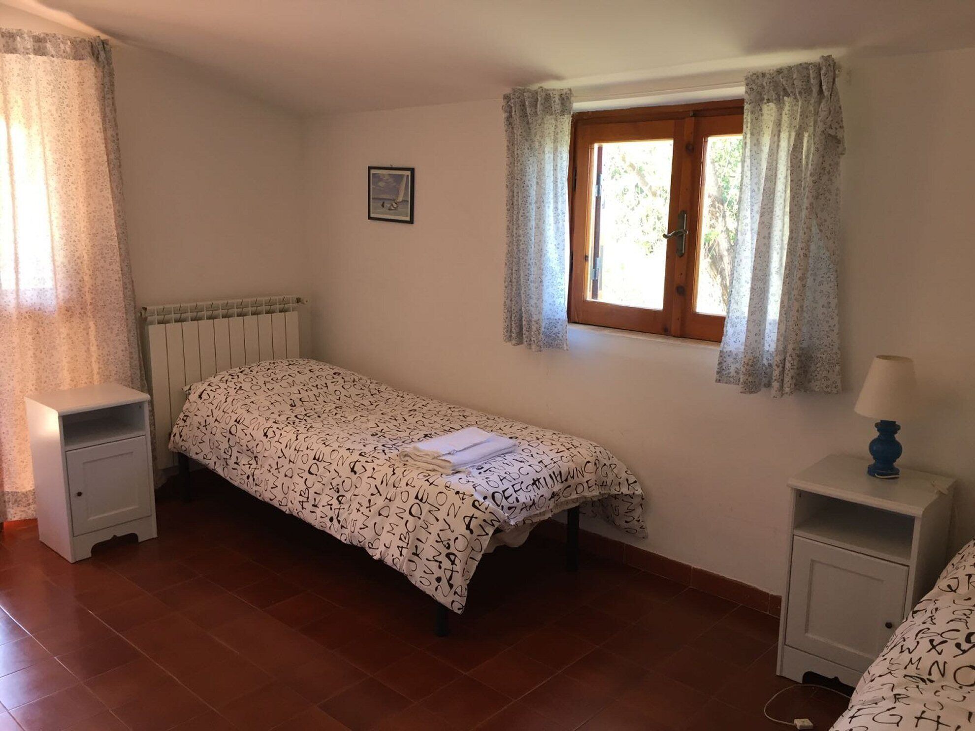 Ein kleines, ordentliches Schlafzimmer mit einem Einzelbett, einem Nachttisch, einer Fensterbank, Vorhängen und einer Heizung.