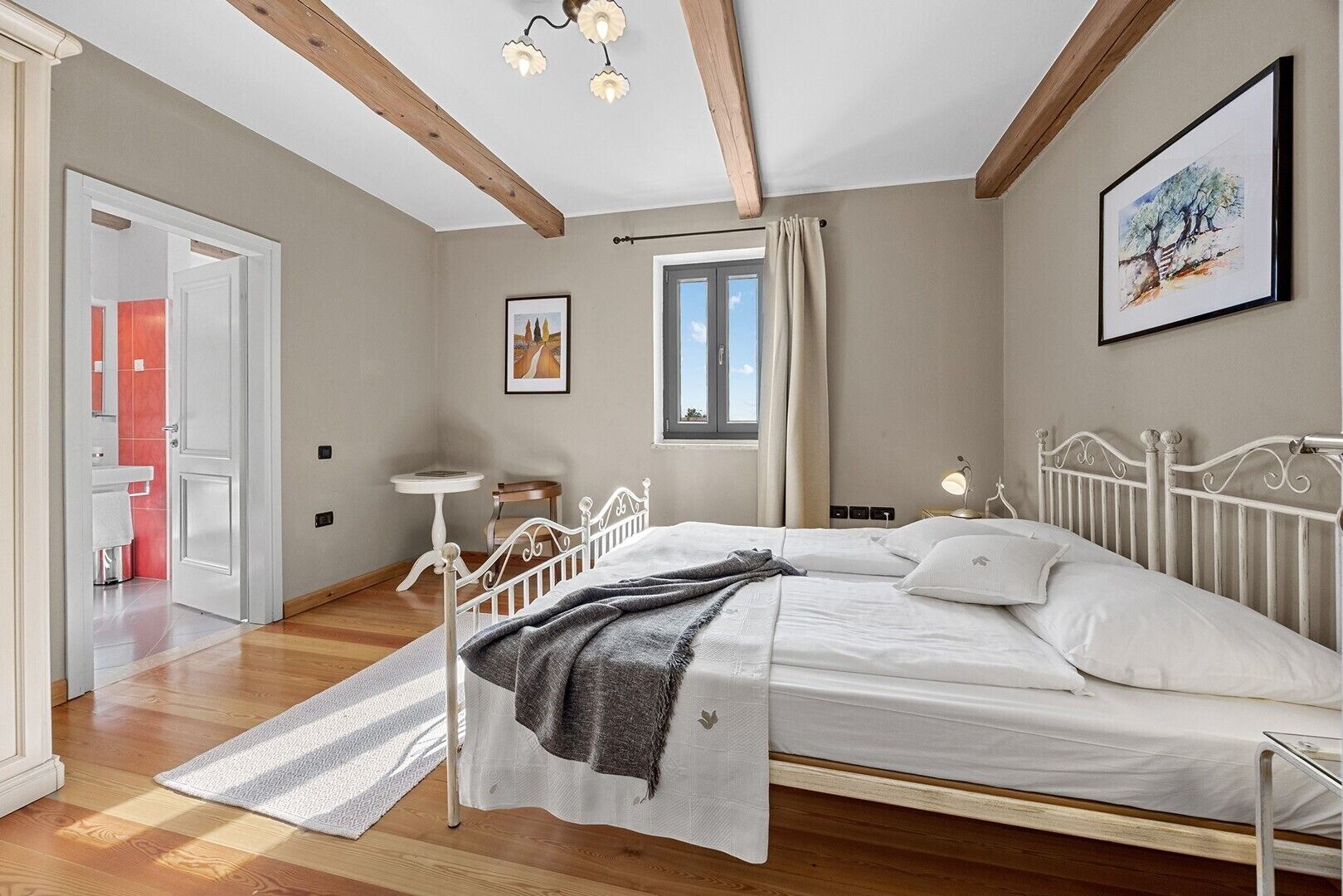 A bedroom with a bed, a bedside table, a window with curtains, a door to another room, and a painting on the wall.