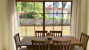 Un espace repas comprenant une table et des chaises, de grandes fenêtres avec des rideaux, et une vue sur un jardin et des bâtiments.