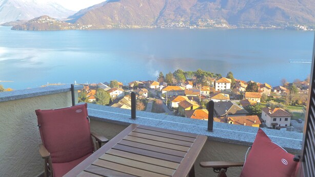 A balcony with a wooden table and two red chairs, overlooking a lake and a village with houses and a mountain in the background.