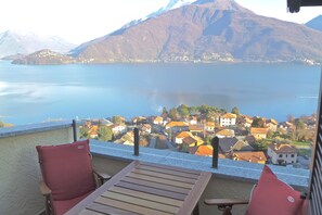 Un balcón con una mesa de madera y dos sillas rojas, con vista a un lago y un pueblo con casas, y una montaña al fondo.