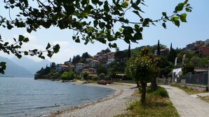 A lakeside view with a pebble beach, a walking path, and buildings in the background.