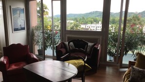 A living room with a red armchair, a dark wooden table, and a sofa with a yellow cushion. Large windows offer a view of greenery and buildings. - Pau facing the Pyrenees (Pau)