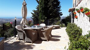 Une terrasse extérieure avec des meubles en osier, une table et un parasol. On y trouve des plantes en pot et une vue sur le paysage.