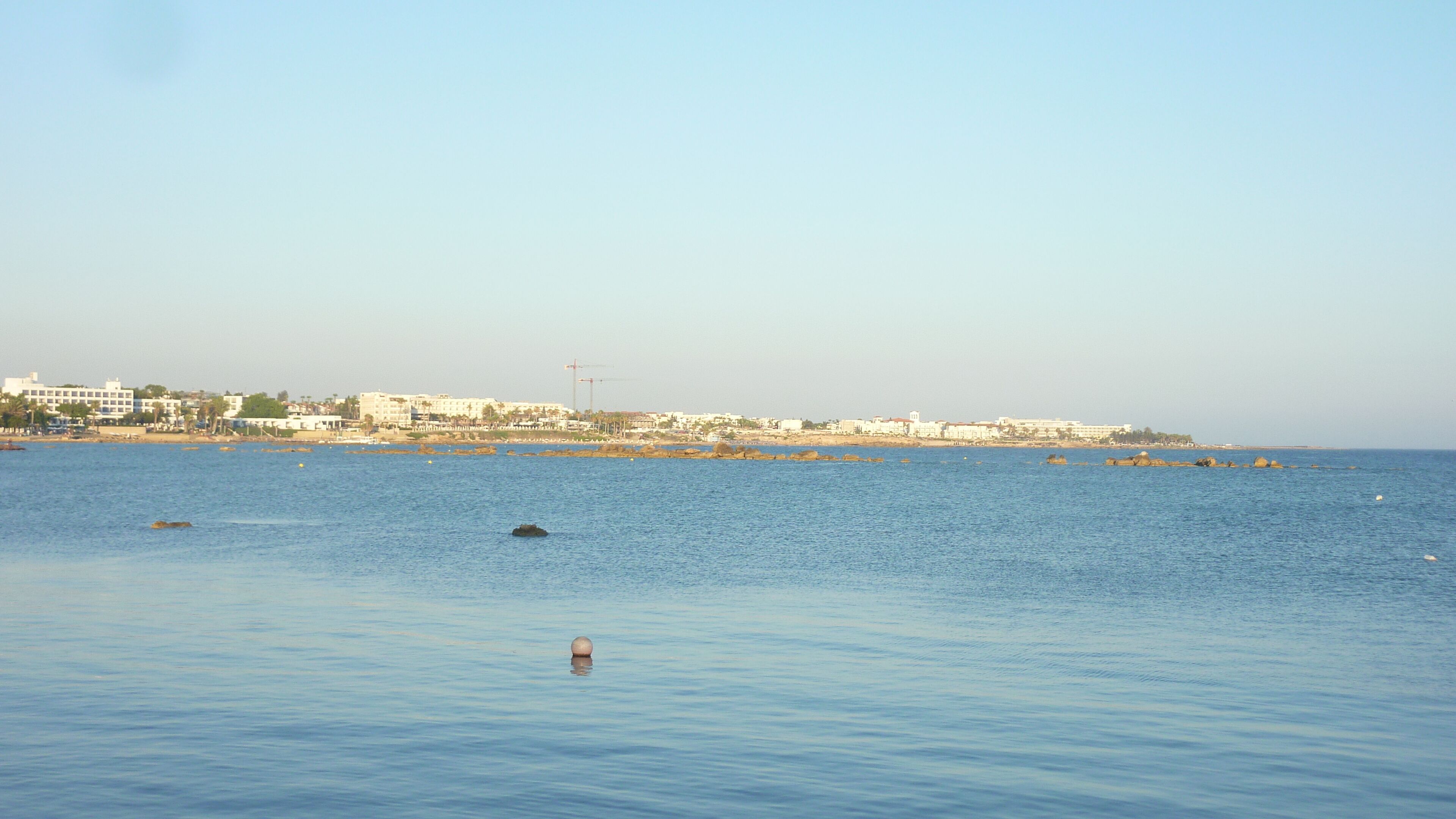 A coastal view with calm waters, a distant shoreline with buildings, and a clear sky.