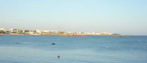 A coastal view with calm waters, a distant shoreline with buildings, and a clear sky.