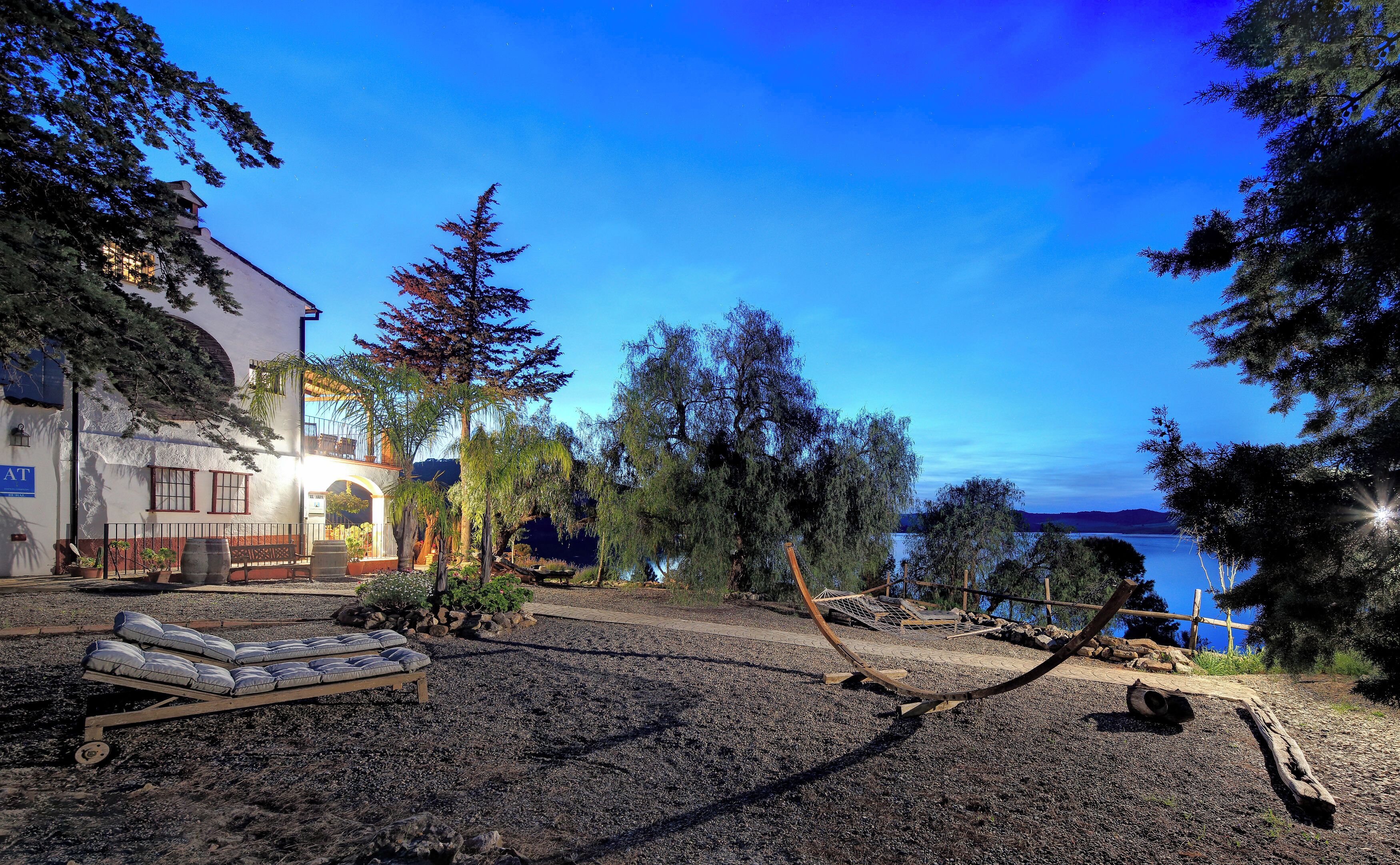A building with a balcony, a gravel area with a bench, and a view of a body of water.