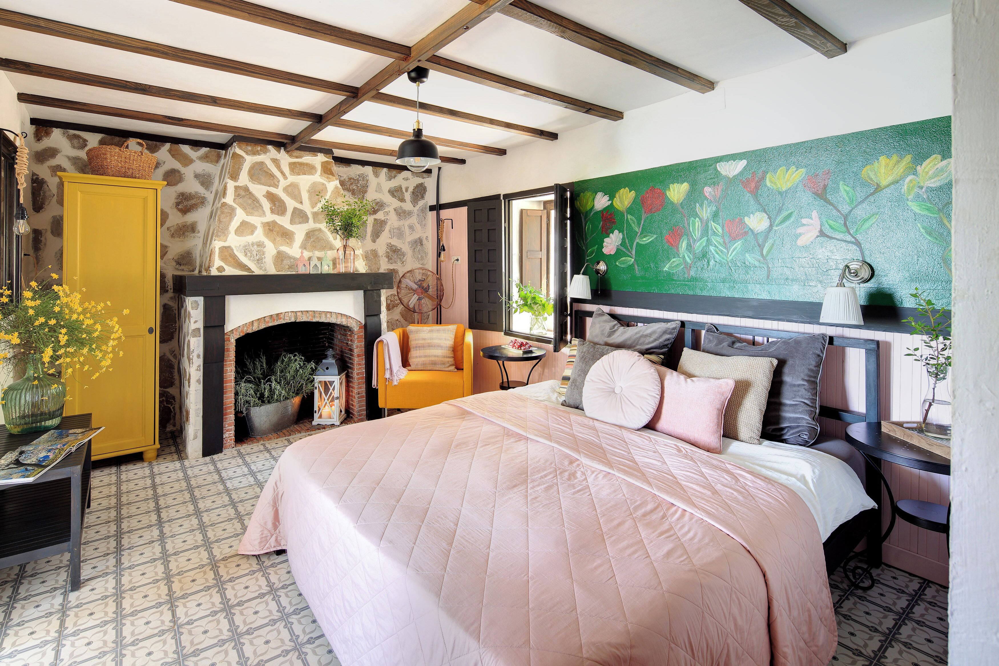 Ein Schlafzimmer mit Bett, Kamin, gelbem Sessel, Tisch mit Lampe und Wand mit floralem Wandbild.