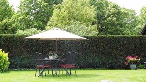 Eine Terrasse mit Tisch, Stühlen und einem Sonnenschirm, im Hintergrund ein Hecken.