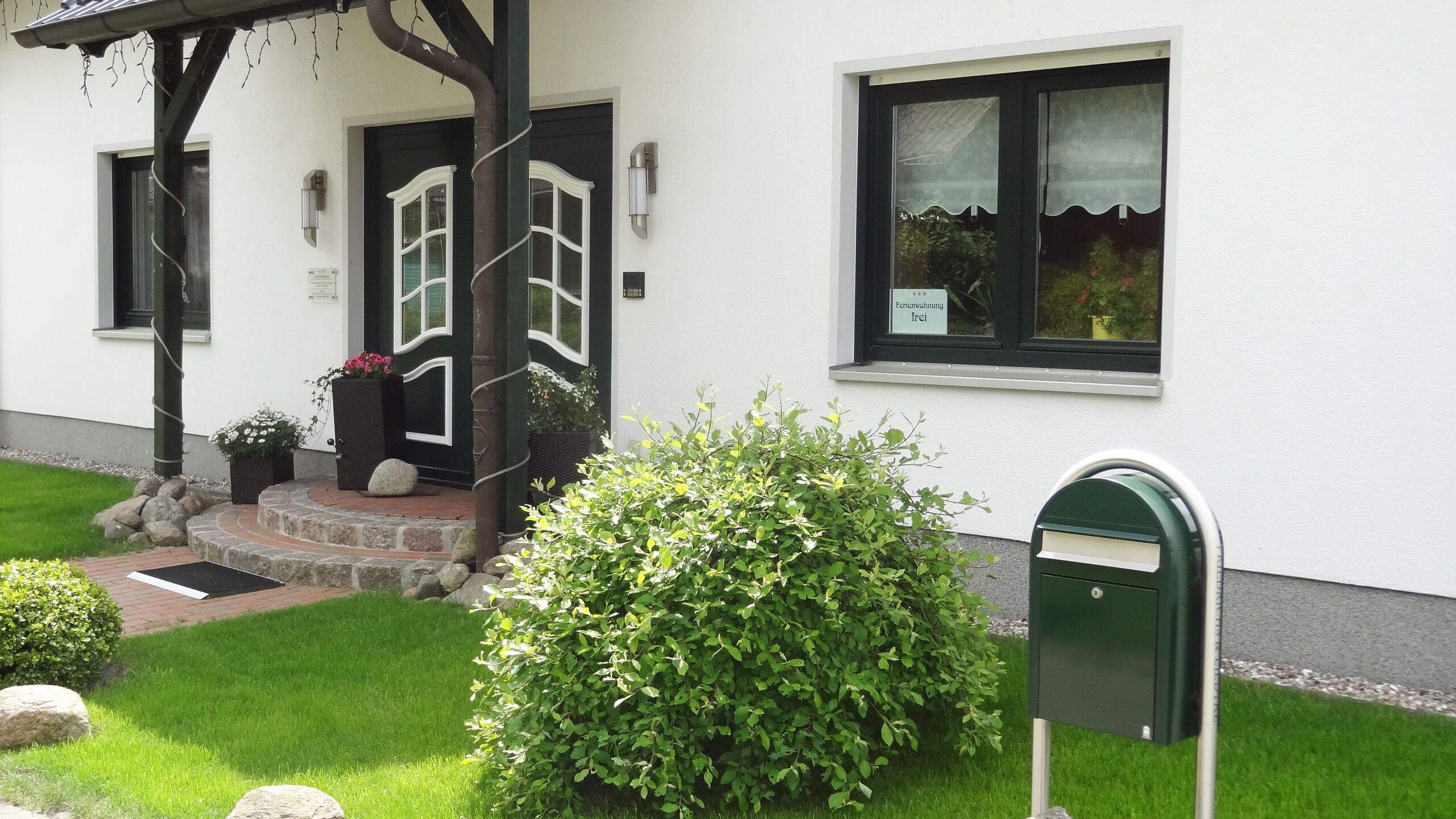 A building with a white exterior, a black door with a white frame, a small porch with steps, a green mailbox, and a window with a curtain.