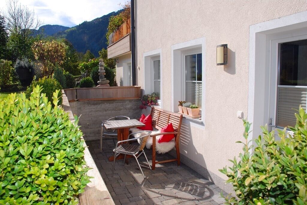 Un patio con una mesa y sillas, un banco de madera con cojines rojos y plantas en macetas.