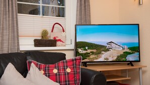 A living room with a television displaying a scenic view, a black leather sofa, and a window with curtains. - 55m² large apartment with terrace and garden and separate entrance (Hippach)