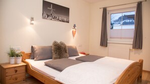 A bedroom with a wooden bed, two bedside tables, a nightstand, a wall-mounted light, a window with curtains, and a painting on the wall. - 55m² large apartment with terrace and garden and separate entrance (Hippach)