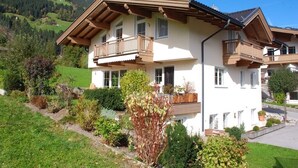 A two-story chalet with wooden balconies, surrounded by lush greenery and a mountainous backdrop. - 55m² large apartment with terrace and garden and separate entrance (Hippach)