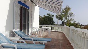 A covered outdoor seating area with white chairs and a table, a white building with a blue stripe, and a tiled floor.