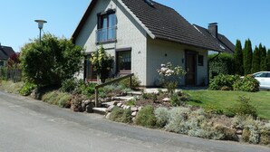 Ein zweistöckiges Haus mit einem geneigten Dach, einer Veranda mit Stufen und einem kleinen Garten mit verschiedenen Pflanzen und Blumen.