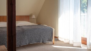 Ein Schlafzimmer mit schrägen Wänden, einem Bett mit grauer Decke, einer Nachttischlampe und einem Fenster mit transparenten Vorhängen.