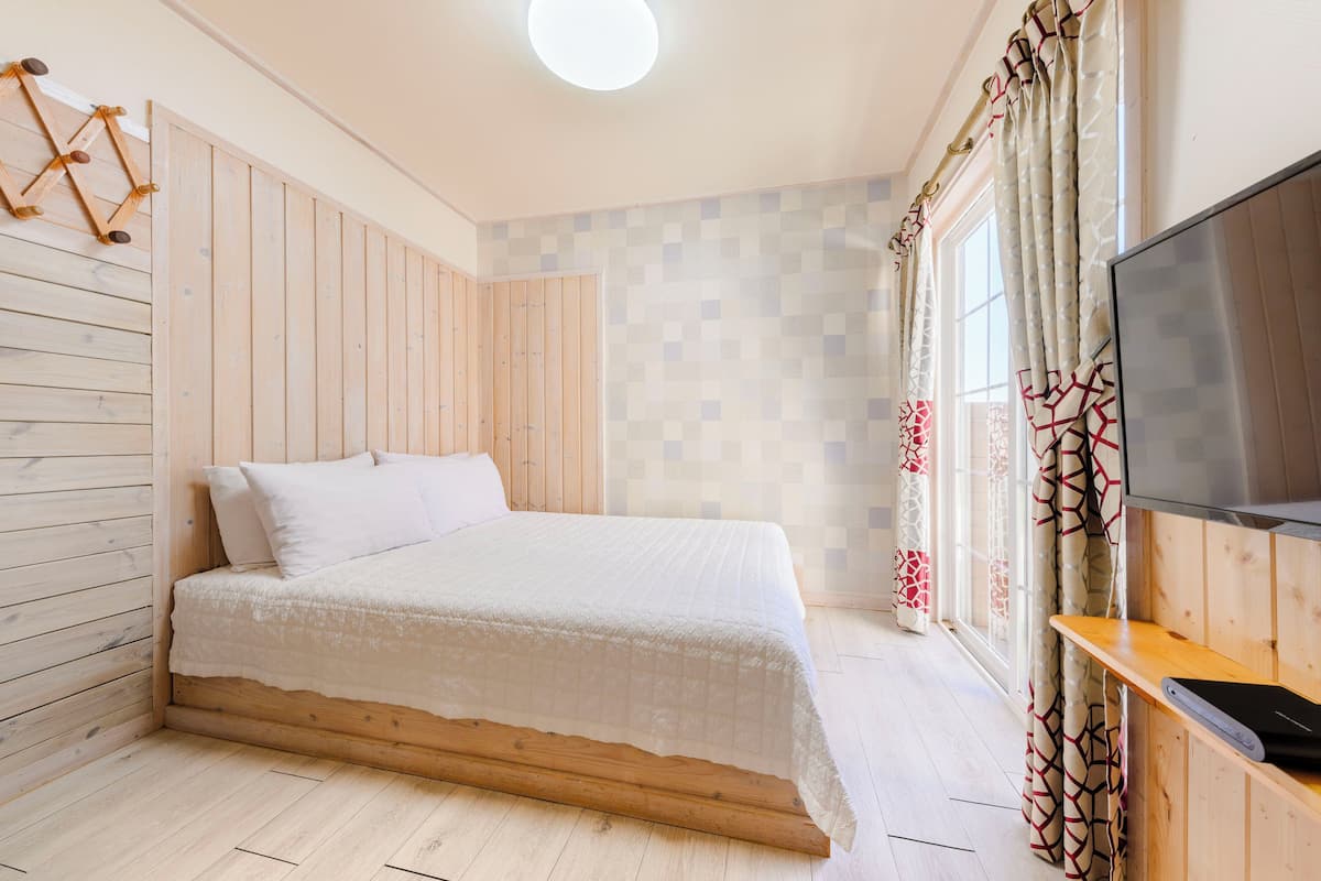 A bedroom with a wooden bed, a TV on a wooden stand, and a window with patterned curtains.