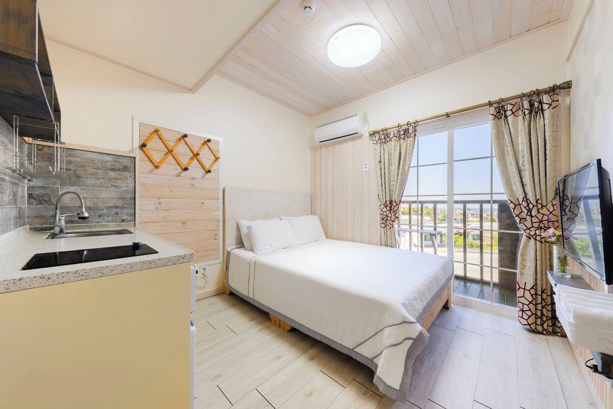 A bedroom with a bed, a window with curtains, a TV, and a kitchen area with a sink and cabinets.