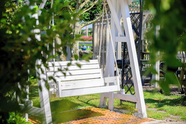 A white swing set with a bench, surrounded by greenery and a brick pathway.