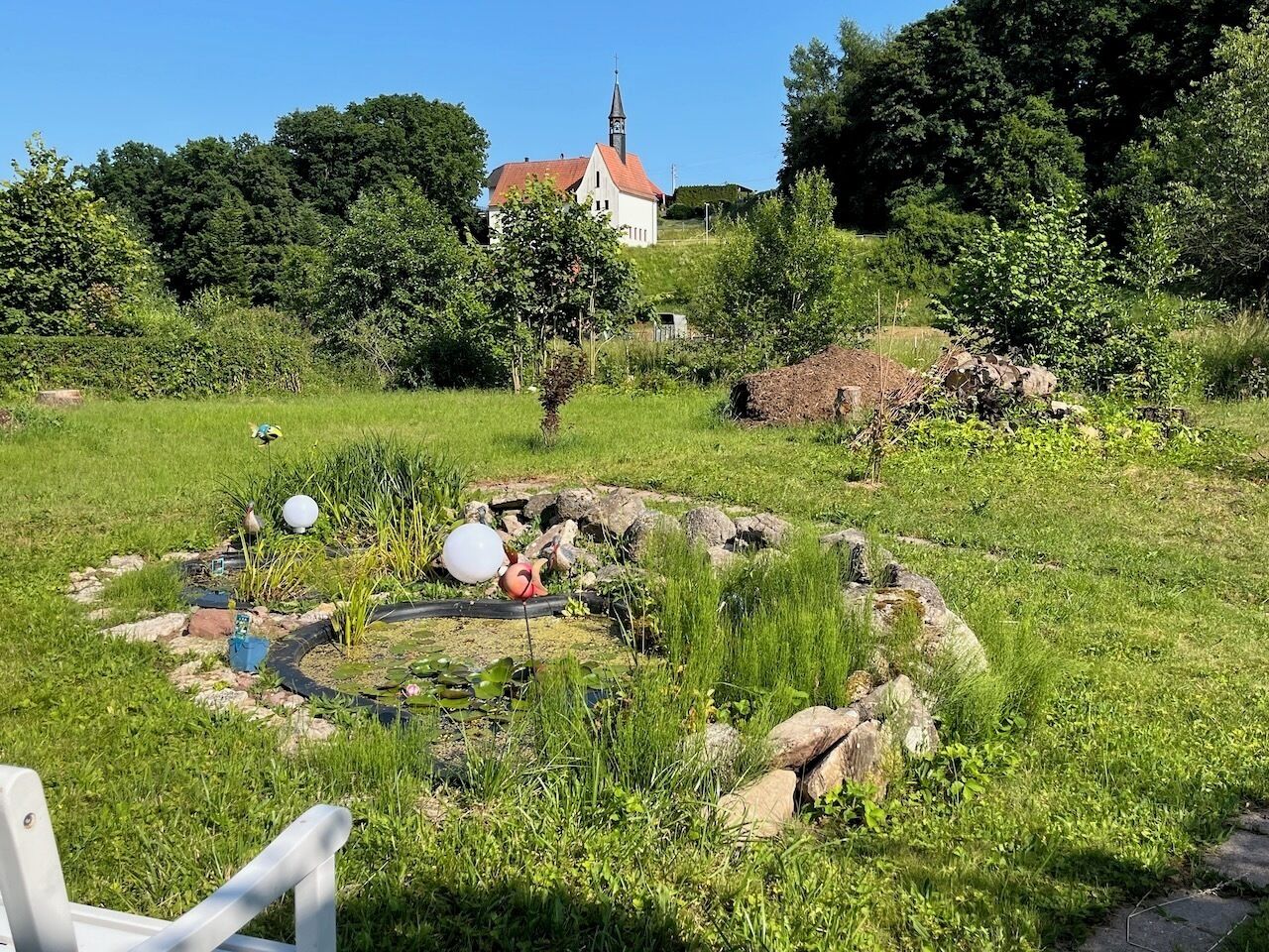 A rural landscape with a white church, a pond with rocks and plants, and a chair in the foreground.