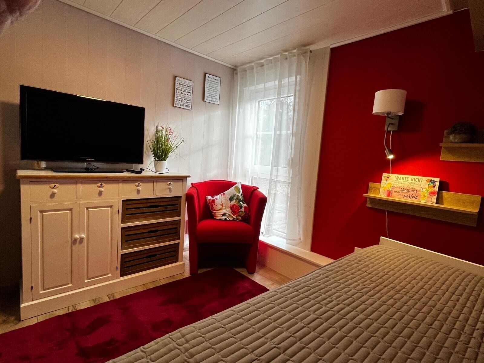 Habitación de hotel con una televisión sobre un mueble, una silla roja, una cama, una ventana con cortinas de gasa y una lámpara fijada a la pared.