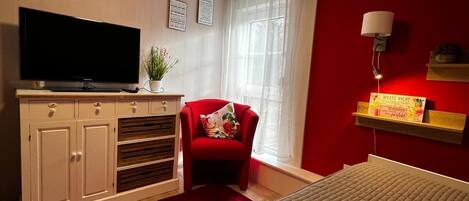 Habitación de hotel con una televisión sobre un mueble, una silla roja, una cama, una ventana con cortinas de gasa y una lámpara fijada a la pared.