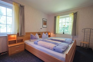 Ein Doppelbett mit Kopfteil, zwei Nachttische, ein kleiner Hocker und ein Fenster mit Vorhängen.