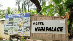 A sign for Hotel Nehapalace with a wooden fence and tropical plants in the background.