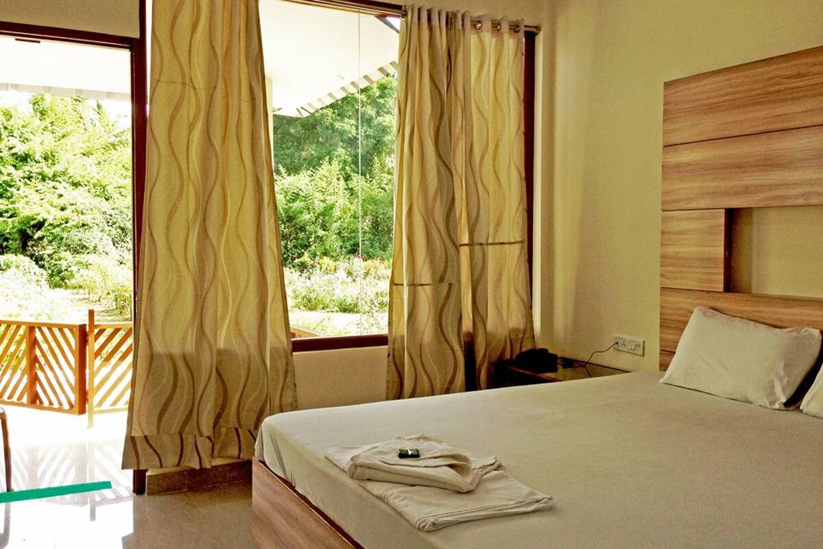 Quarto com cama, cortinas, cabeceira de madeira e vista para o verde lá fora.