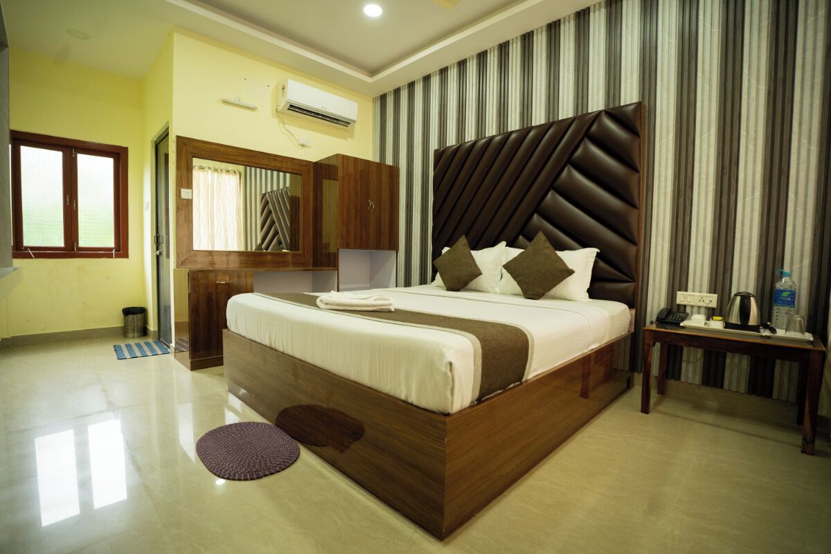 A hotel room with a large bed, a wooden headboard, a small bedside table with a lamp, and a window with curtains.