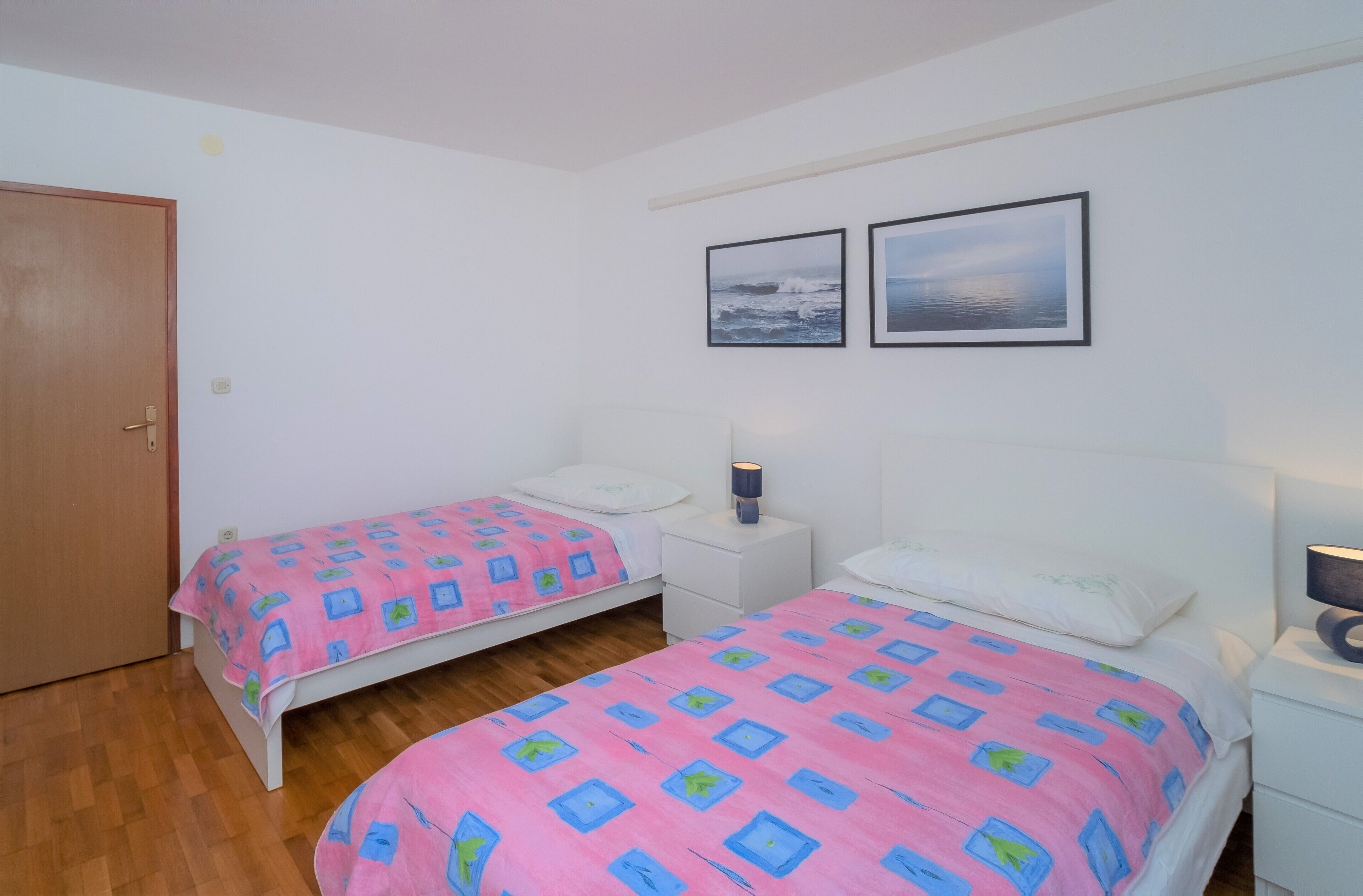Ein Schlafzimmer mit zwei Einzelbetten, weißen Nachttischen, einer Holztür und zwei gerahmten Bildern an der Wand.