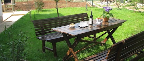 An outdoor patio with a wooden table and benches, surrounded by a well-maintained lawn and a brick oven.