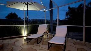 A covered outdoor seating area with two lounge chairs and a large umbrella, overlooking a residential area. - HD Smart TV's, Kayaks, Pool & Spa, WIFI, Gulf Access, E-Dart Board, Bar, Grill  (Cape Coral)