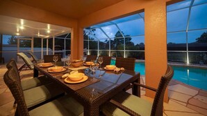 An outdoor dining area with a table set for four, surrounded by wicker chairs with cushions. Large windows provide a view of a swimming pool and trees. - HD Smart TV's, Kayaks, Pool & Spa, WIFI, Gulf Access, E-Dart Board, Bar, Grill  (Cape Coral)