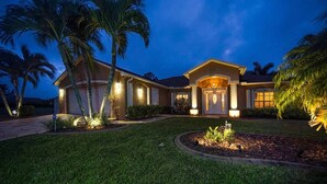 A two-story house with a well-lit front entrance, a garage, and a landscaped front yard with palm trees. - HD Smart TV's, Kayaks, Pool & Spa, WIFI, Gulf Access, E-Dart Board, Bar, Grill  (Cape Coral)