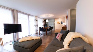 A modern living room with a flat-screen TV, grey sofas, a wooden dining table, and a kitchen in the background. - 6 min from the center and the MatterhornParadise station, next to the ski bus stop (Zermatt)
