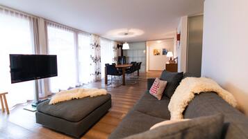A modern living room with a flat-screen TV, grey sofas, a wooden dining table, and a kitchen in the background.
