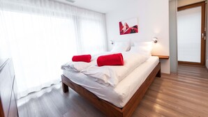 A modern hotel room with a wooden bed, white linens, red towels, a painting on the wall, and a large window with sheer curtains. - 6 min from the center and the MatterhornParadise station, next to the ski bus stop (Zermatt)