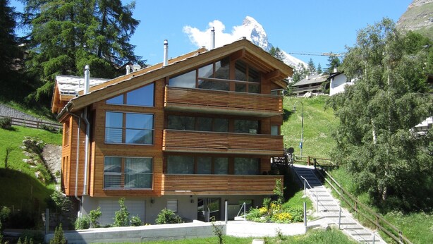 A modern two-story wooden building with large windows and a balcony, surrounded by lush greenery and a mountainous backdrop. - 6 min from the center and the MatterhornParadise station, next to the ski bus stop (Zermatt)