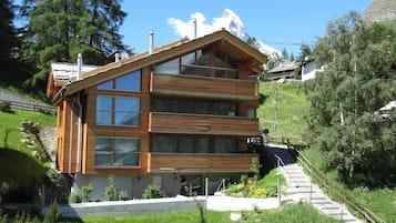 A modern two-story wooden building with large windows and a balcony, surrounded by lush greenery and a mountainous backdrop.