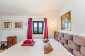 A bedroom with a large bed, a wooden nightstand, a window with a view, and two framed artworks on the wall.