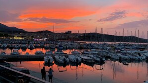 A marina at sunset with boats docked, a cityscape in the background, and a few people by the water.