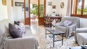A cozy living room with a sofa, armchairs, a coffee table, and a dining table. There is a door leading to an outdoor area with a view of greenery. - Holiday house with pool, air conditioning in the bedrooms, internet / outside kitchen with barbecue, (Pollensa)