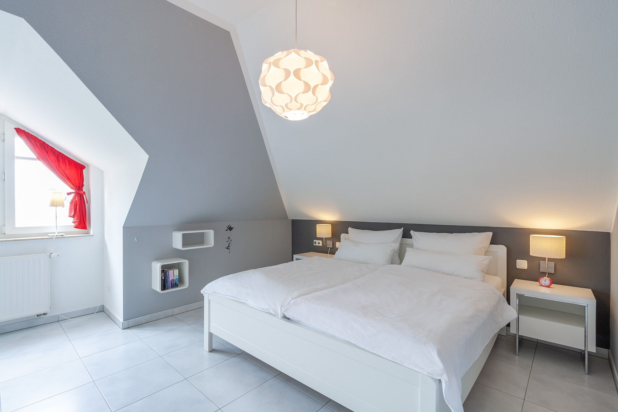 A modern bedroom with a sloped ceiling, a bed with white linens, bedside lamps, and a unique pendant light.